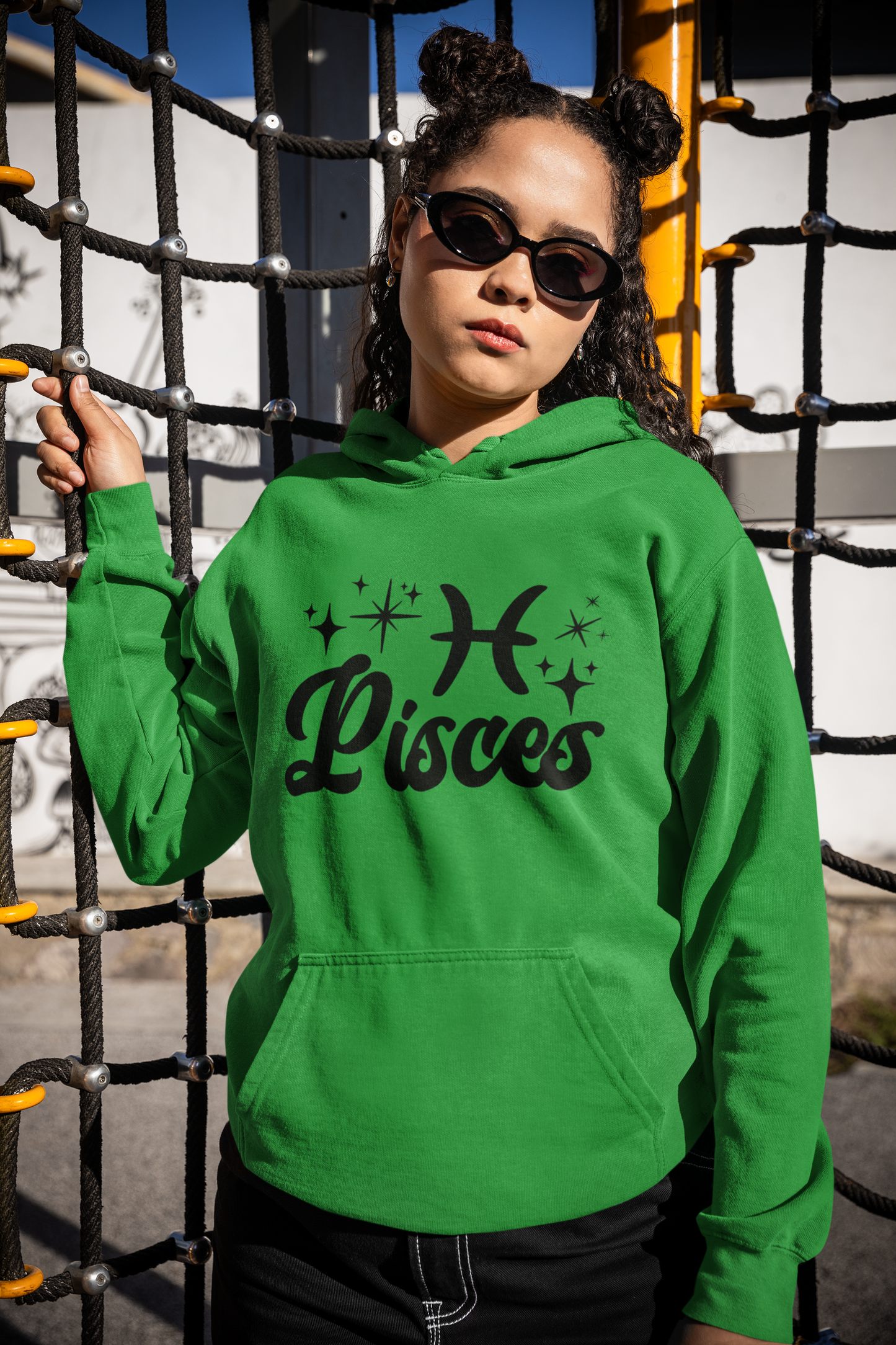 Person wearing a green hoodie with 'Pisces' black text with zodiac symbol, standing in front of playground equipment.