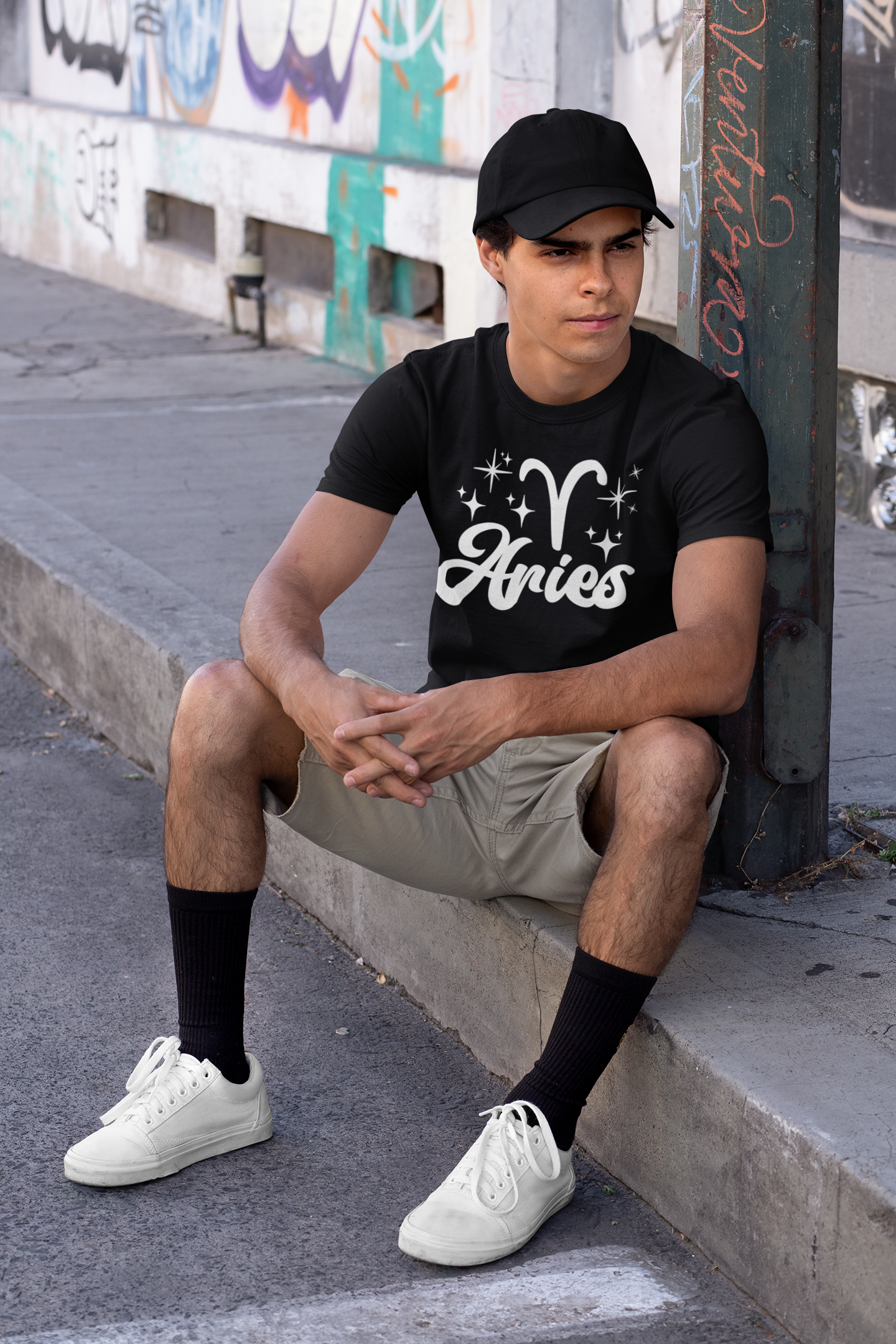 Person wearing a black 'Aries' white text design t-shirt sitting on a sidewalk.