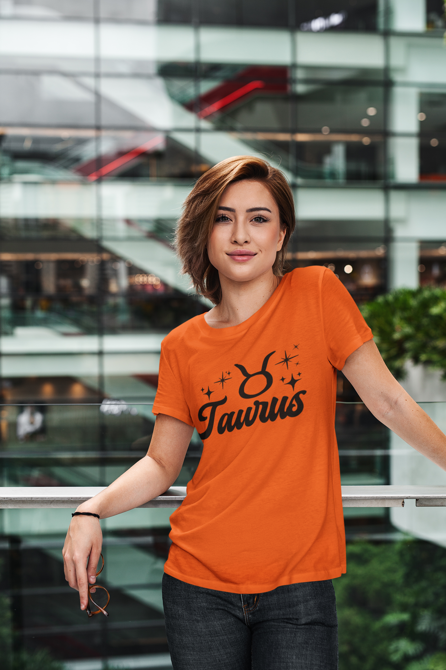 Person wearing a orange Taurus t-shirt, black text design in front of a modern building.
