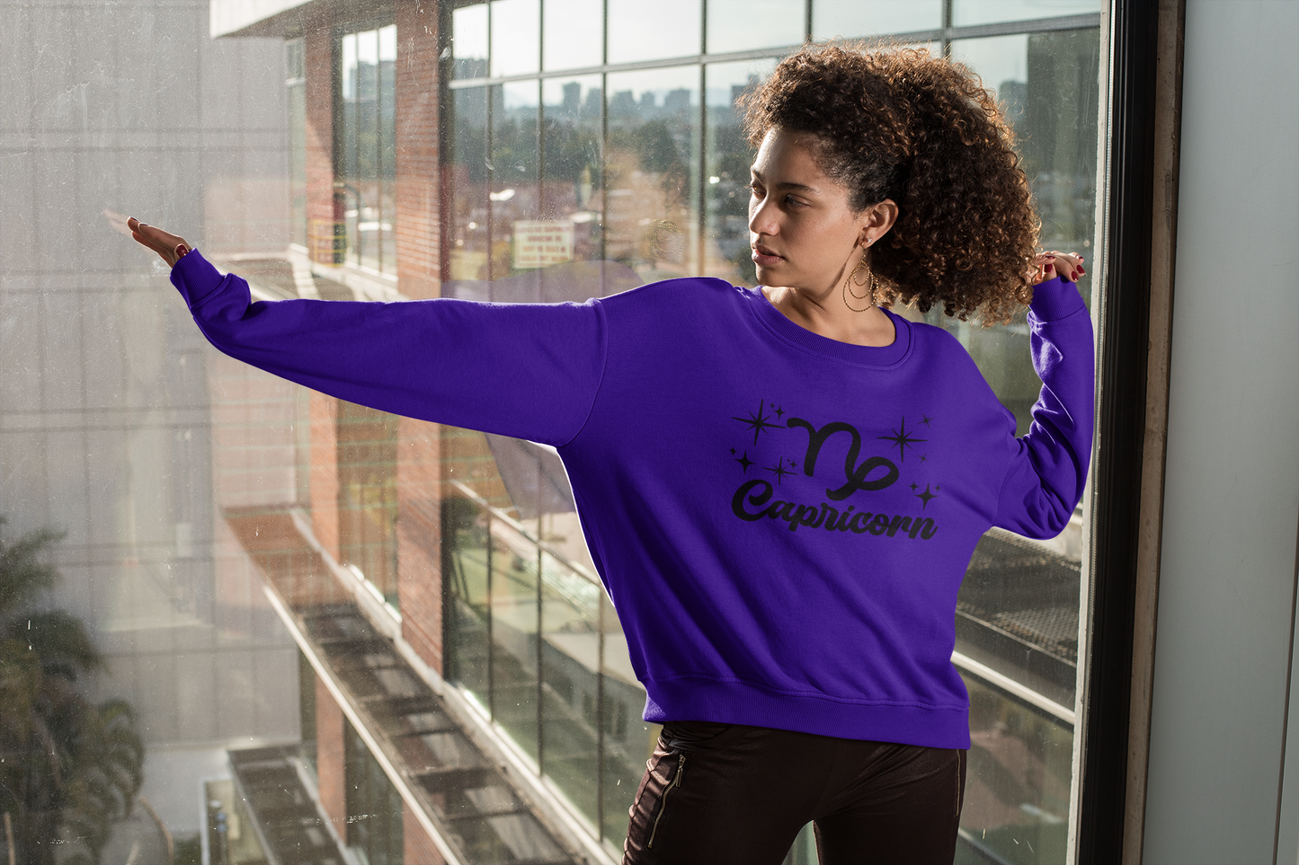 Person wearing a purple 'Capricorn' black text design sweatshirt in front of a window with a cityscape view.