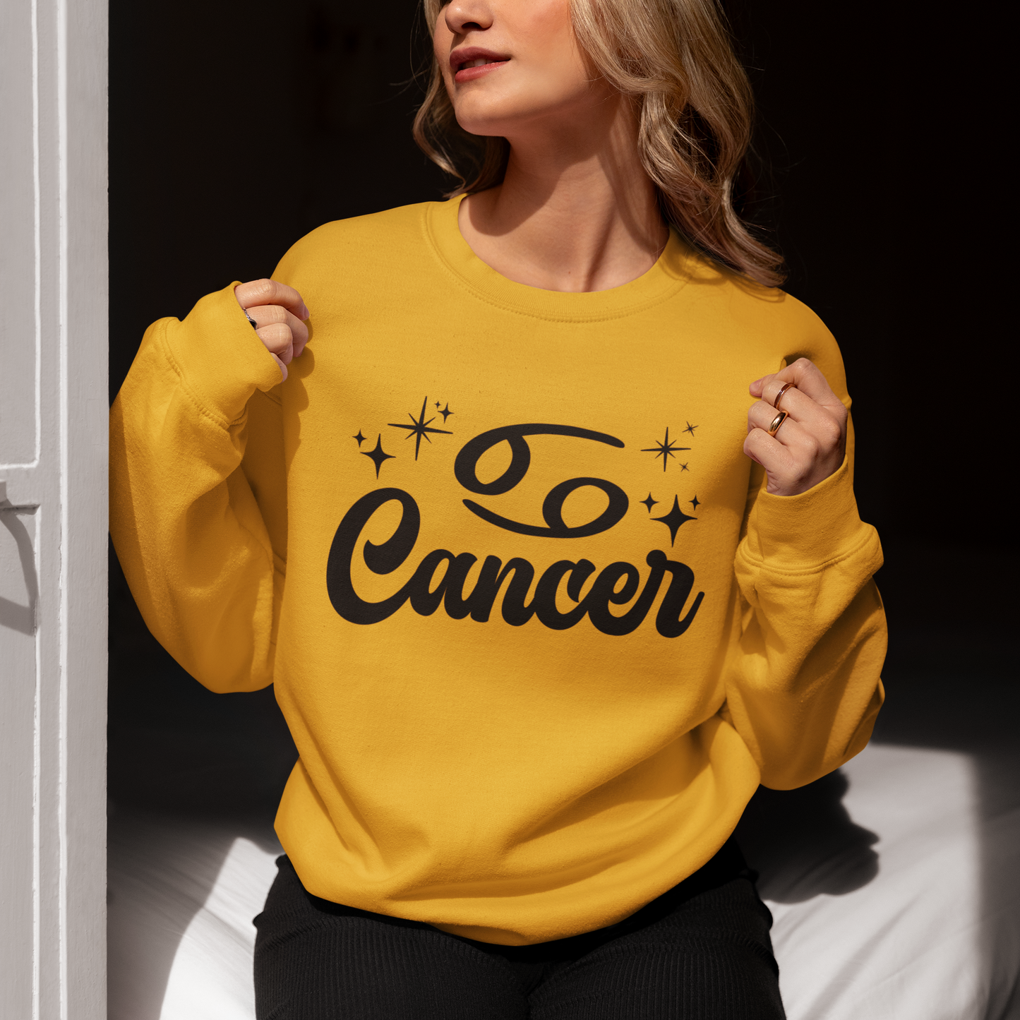 Person wearing a gold sweatshirt with 'Cancer' black text, sitting indoors.