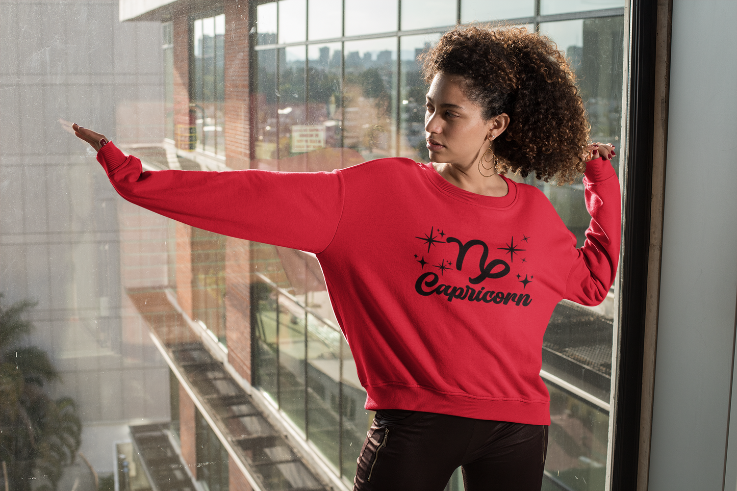 Person wearing a red 'Capricorn' black text design sweatshirt in front of a window with a cityscape view.