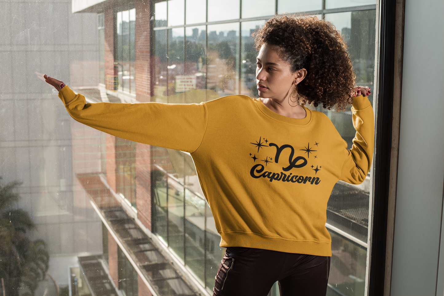 Person wearing a gold 'Capricorn' black text design sweatshirt in front of a window with a cityscape view.