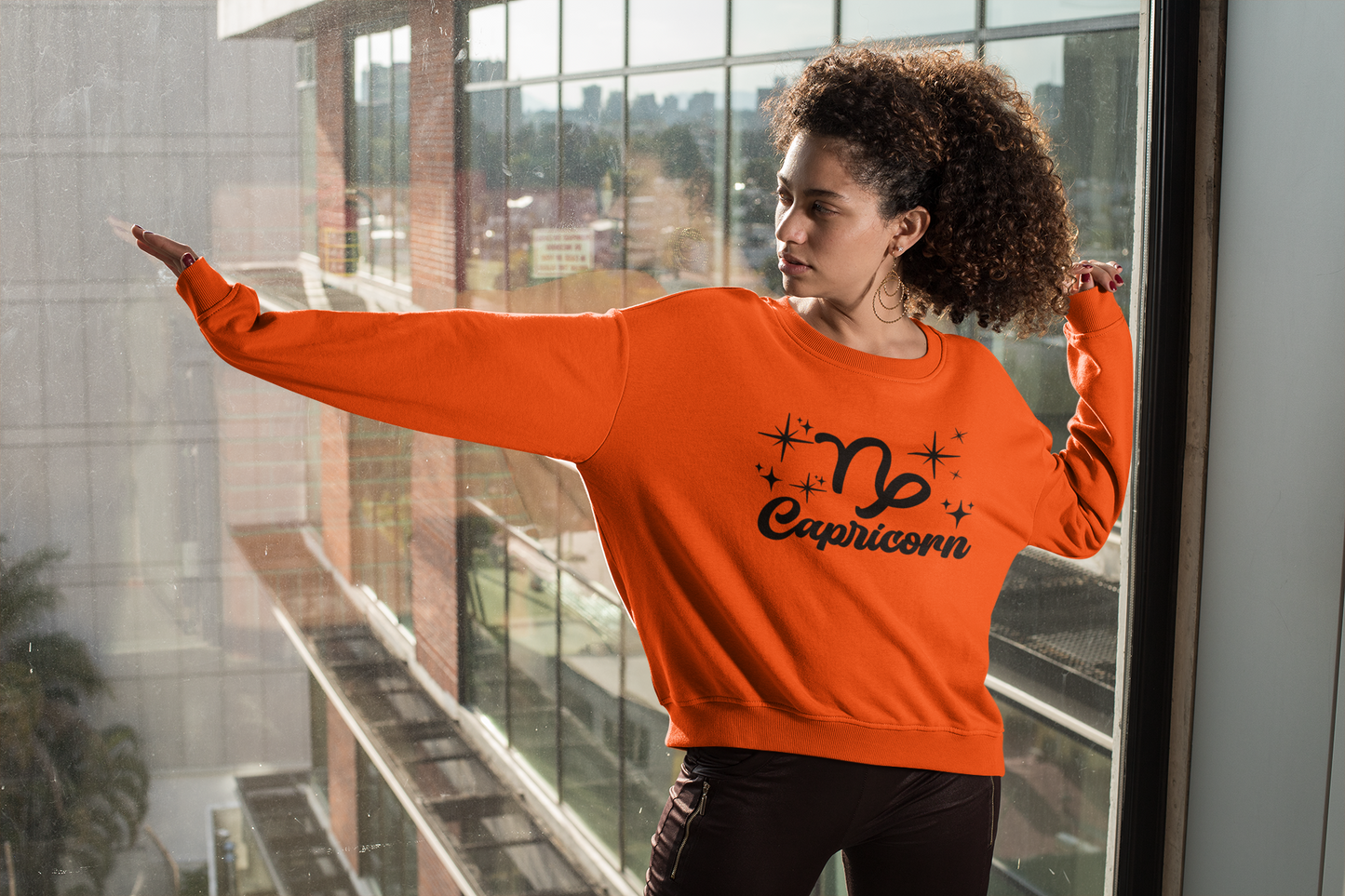 Person wearing a orange 'Capricorn' black text design sweatshirt in front of a window with a cityscape view.