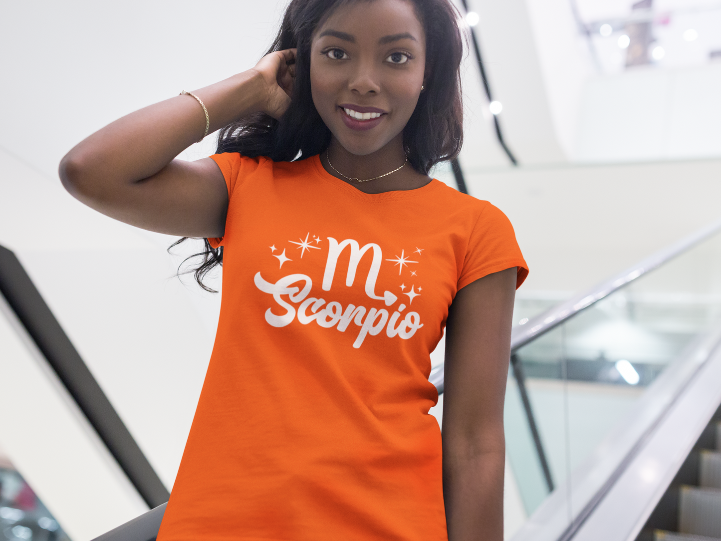 Woman wearing a orange t-shirt with 'Scorpio' white text design, standing on escalator.