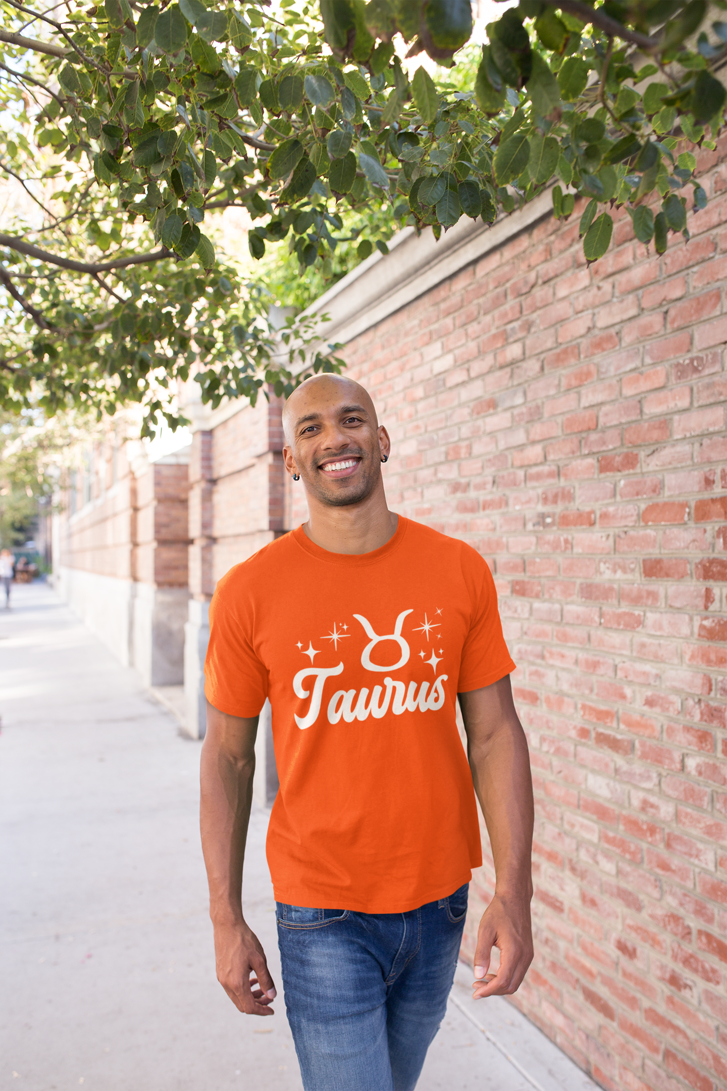 Man wearing a orange t-shirt with 'Taurus' white text design, walking outdoors near a brick wall and tree.