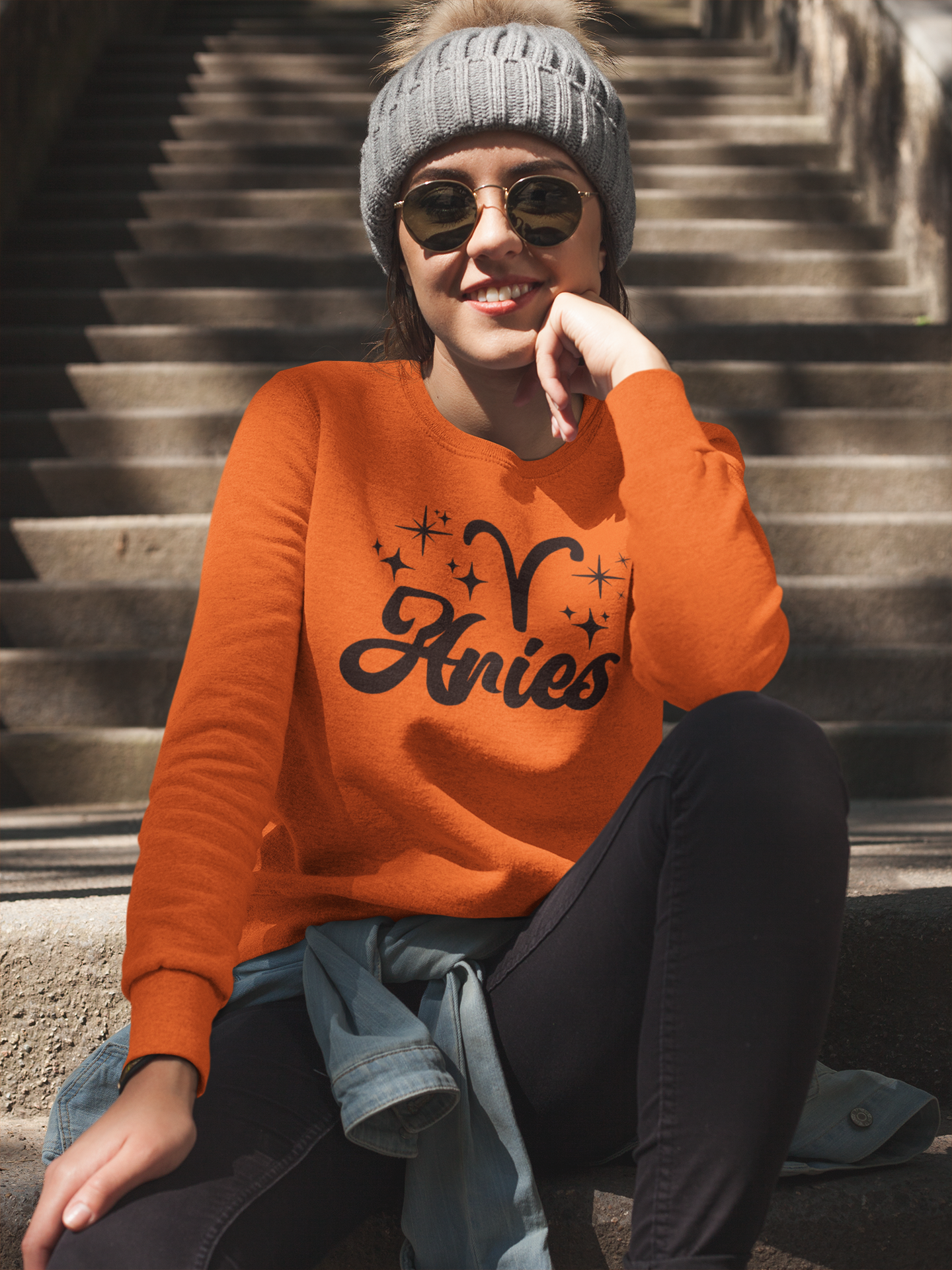 Person wearing a orange 'Aries' black text design sweatshirt sitting on steps.
