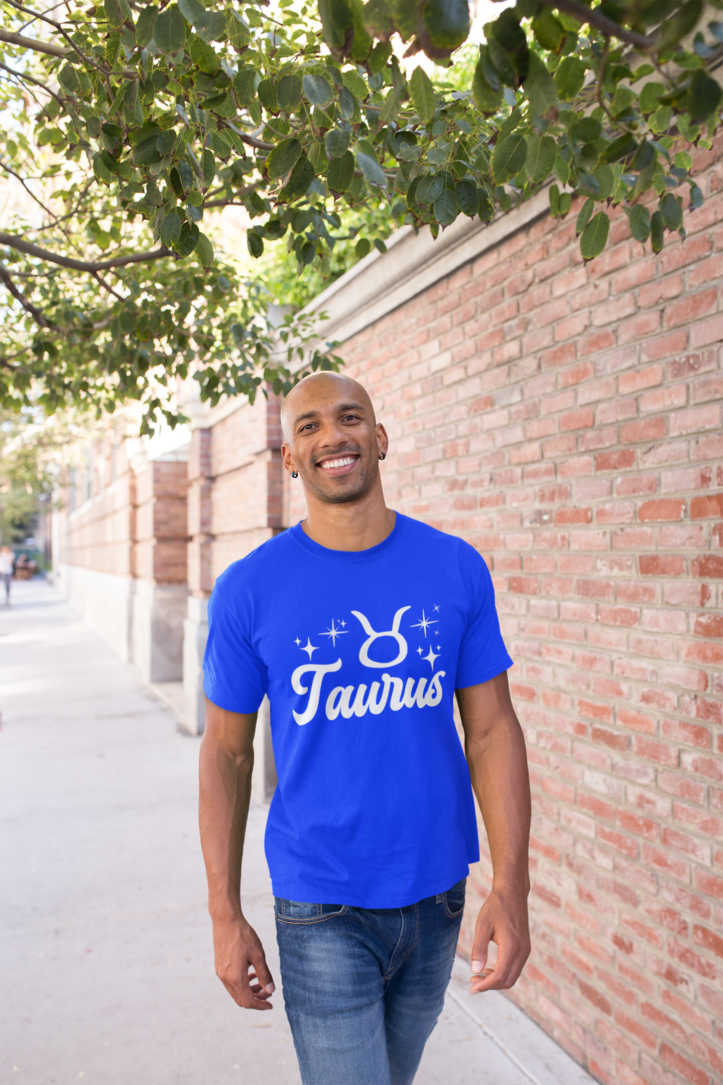 Man wearing a blue t-shirt with 'Taurus' white text design, walking outdoors near a brick wall and tree.