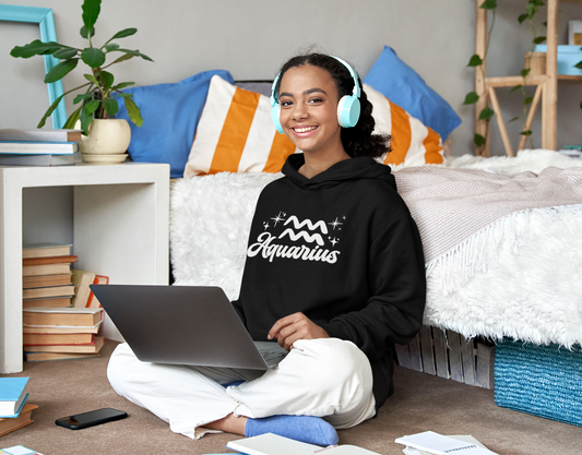 Person wearing a black hoodie 'Aquarius' white text with zodiac symbol, sitting on a bed with a laptop, surrounded by books and a plant.