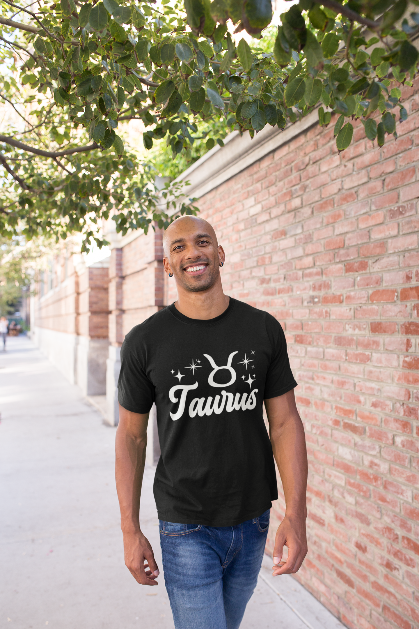 Man wearing a black t-shirt with 'Taurus' white text design, walking outdoors near a brick wall and tree.