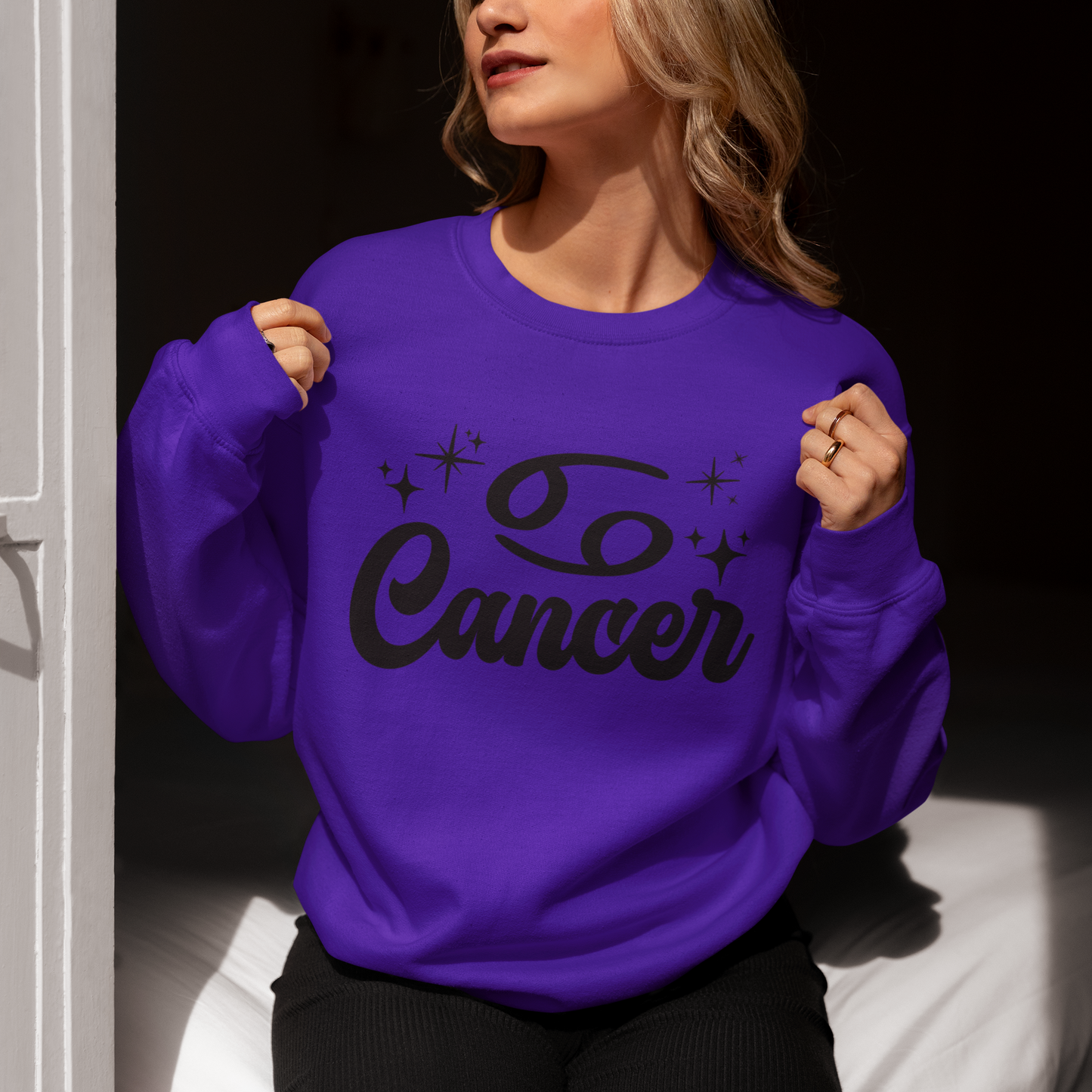 Person wearing a purple sweatshirt with 'Cancer' black text, sitting indoors.