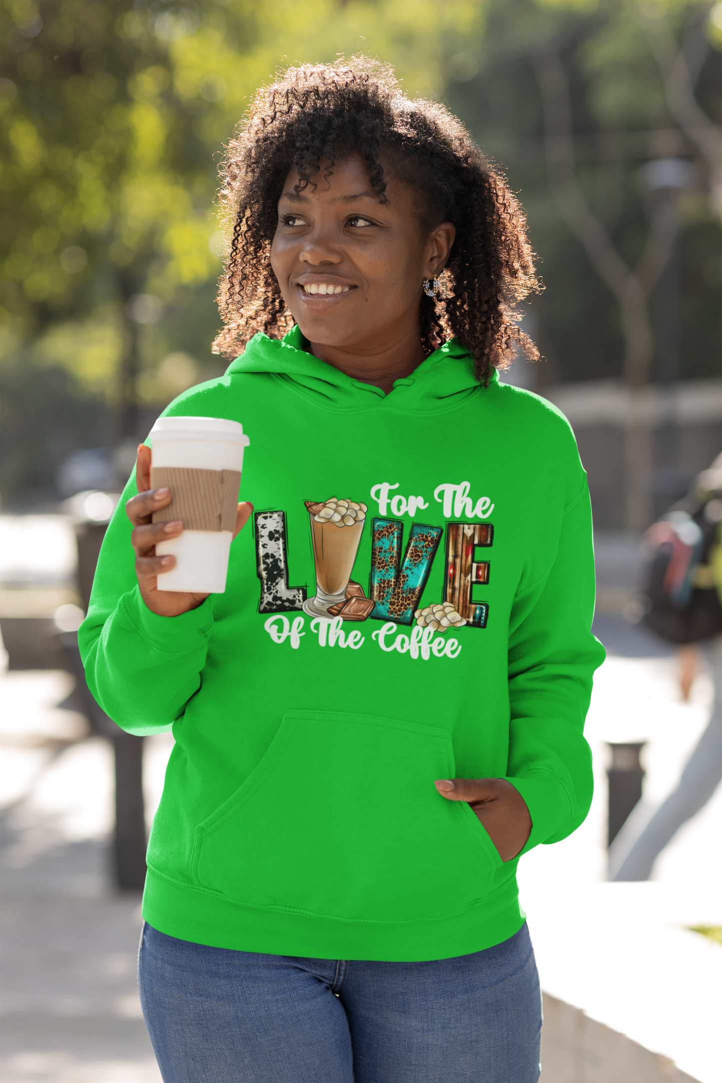 Person wearing a green hoodie with coffee-themed text, holding a coffee cup outdoors.