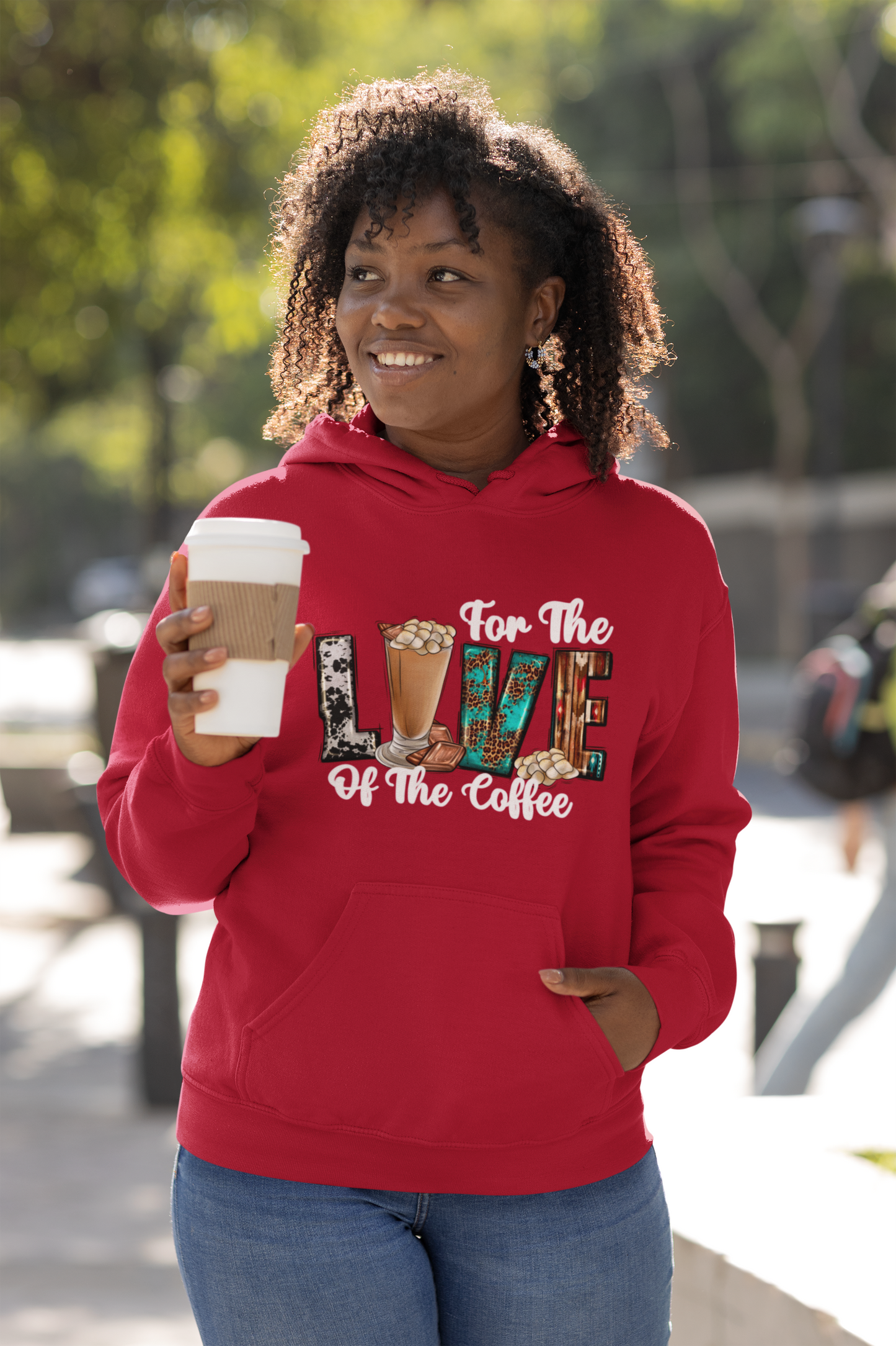Woman wearing a For the love of coffee red hoodie with coffee-themed design, holding a coffee cup outdoors.