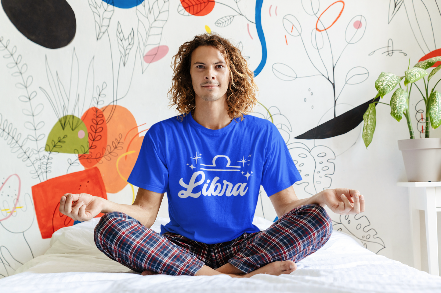 Person wearing an blue 'Libra' white text design t-shirt sitting on a bed with a colorful wall in the background