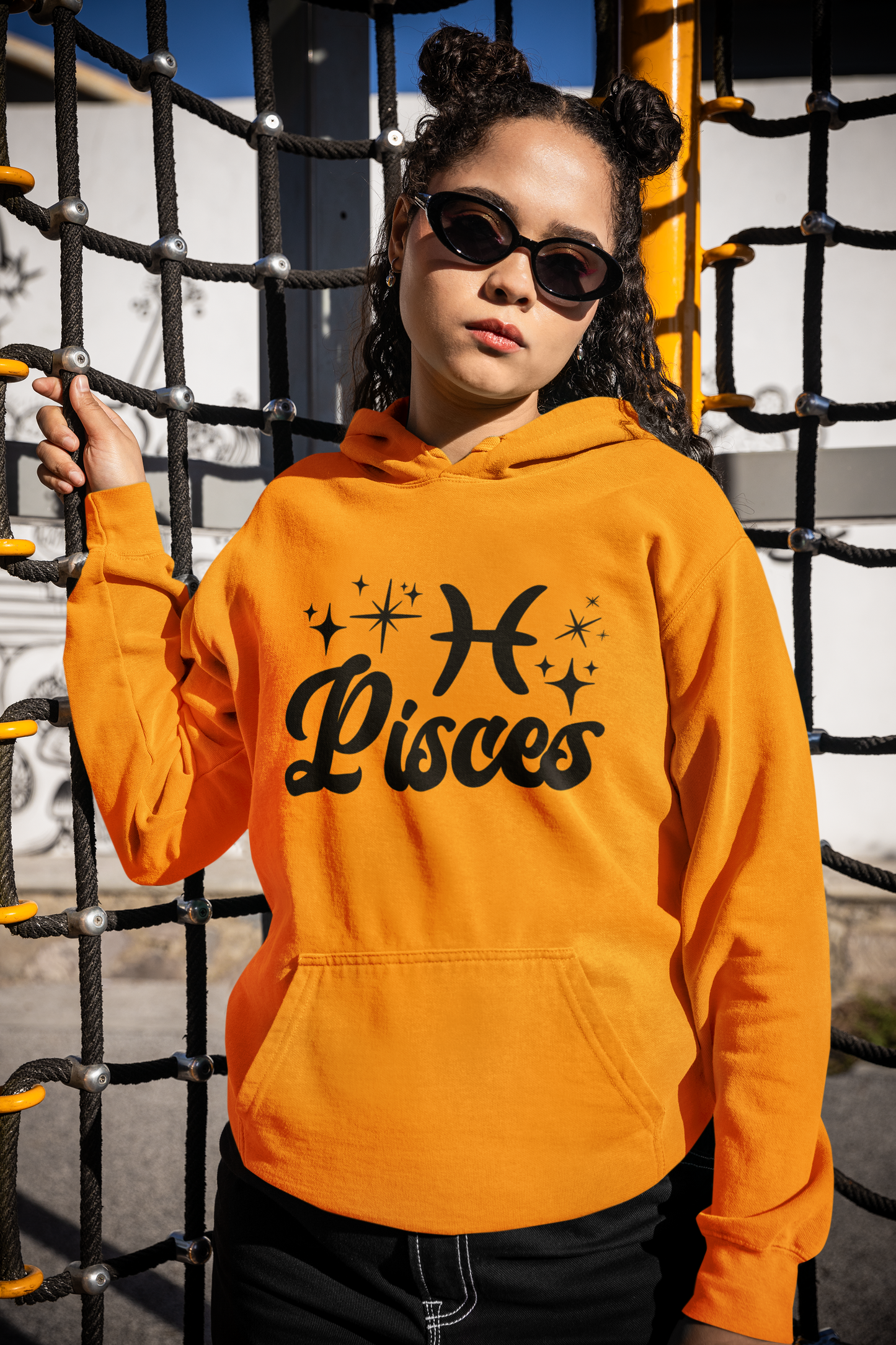 Person wearing a gold hoodie with 'Pisces' black text with zodiac symbol, standing in front of playground equipment.