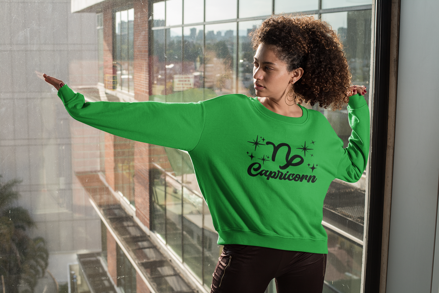 Person wearing a green 'Capricorn' black text design sweatshirt in front of a window with a cityscape view.
