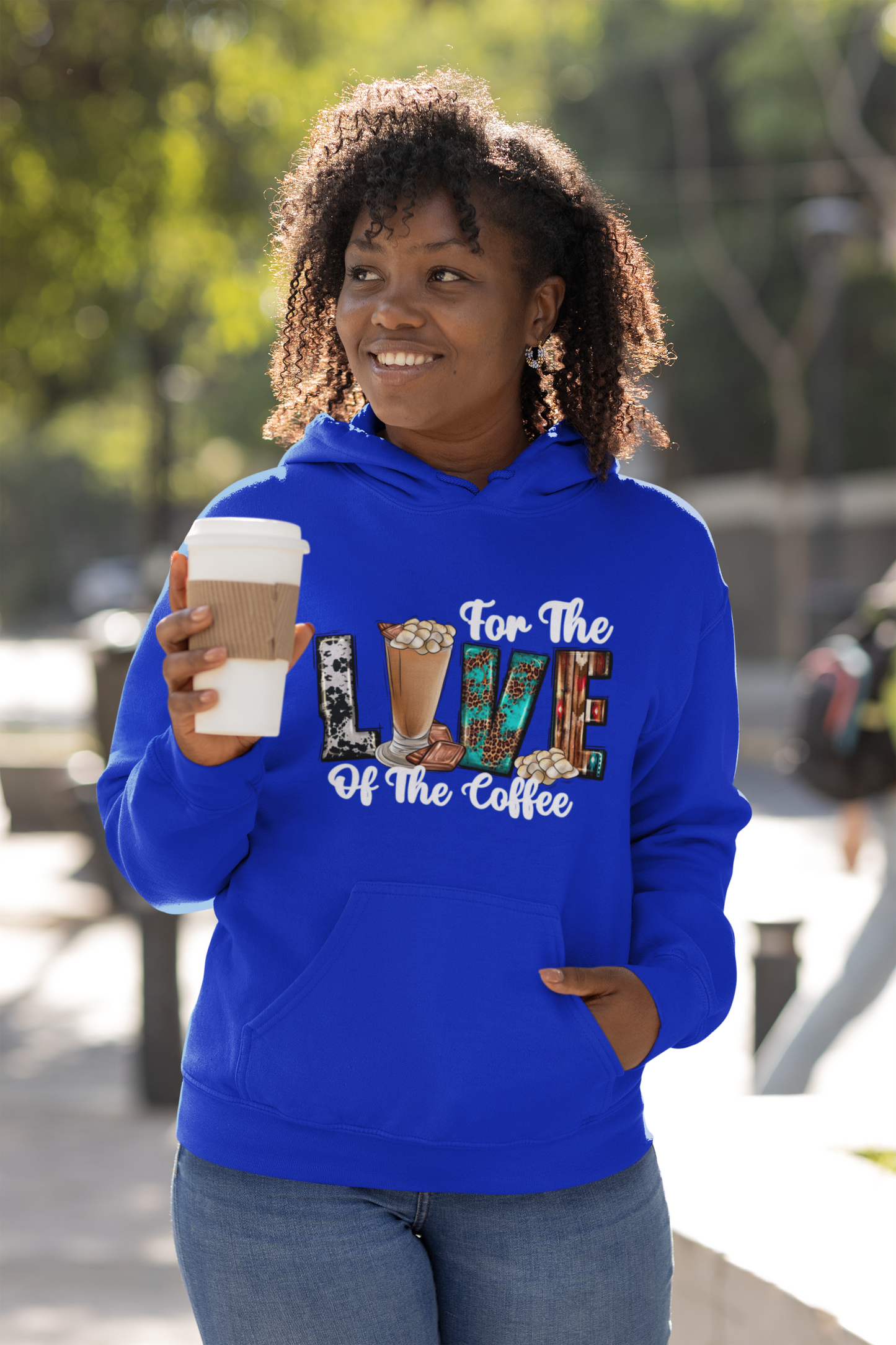 Woman wearing a blue hoodie with coffee-themed text, holding a coffee cup outdoors.