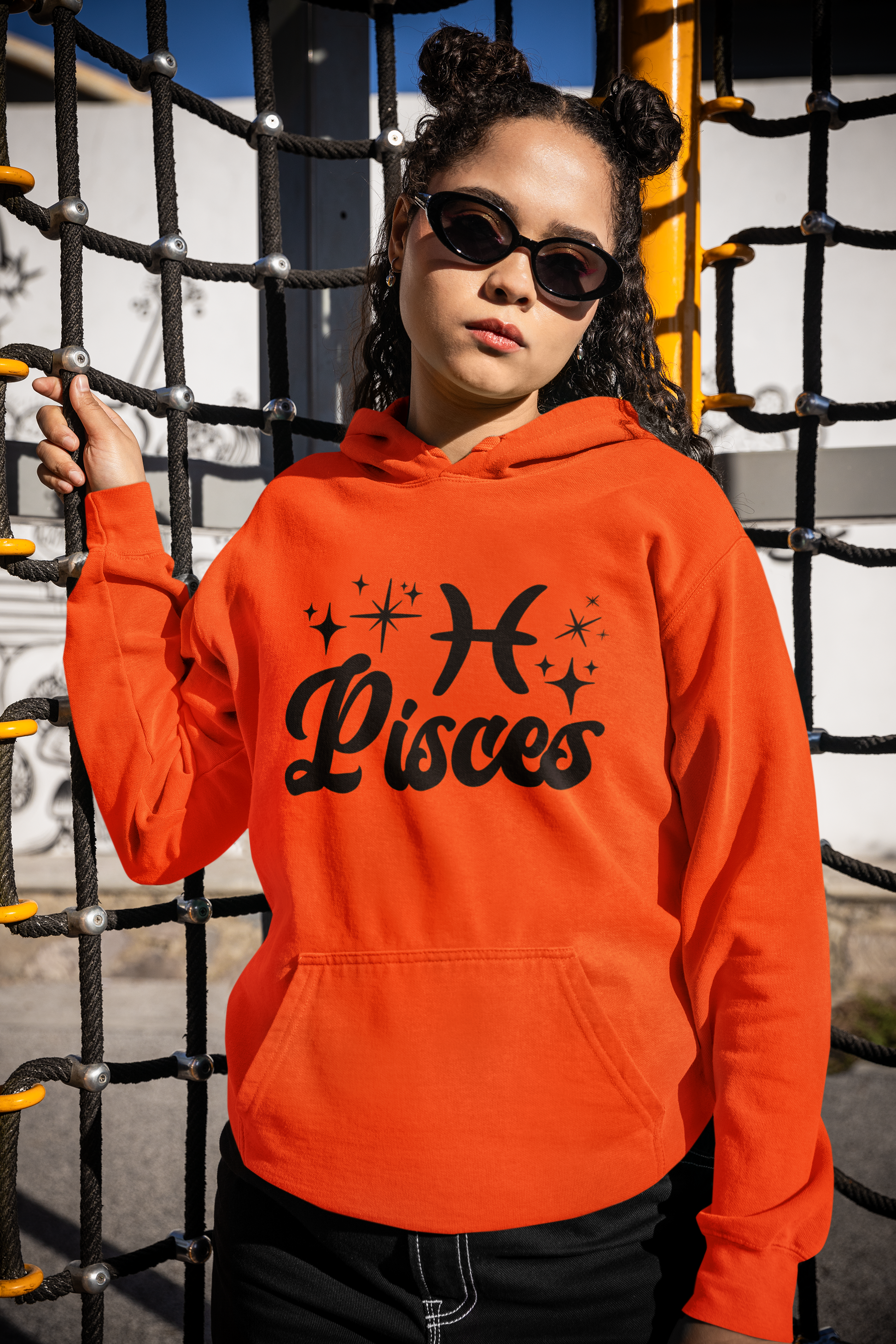 Person wearing a orange hoodie with 'Pisces' black text with zodiac symbol, standing in front of playground equipment.