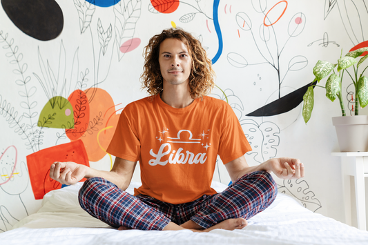 Person wearing an orange 'Libra' white text design t-shirt sitting on a bed with a colorful wall in the background