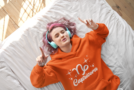 Person wearing an orange 'Capricorn' hoodie - white text design, lying on a bed with pink hair and blue headphones.