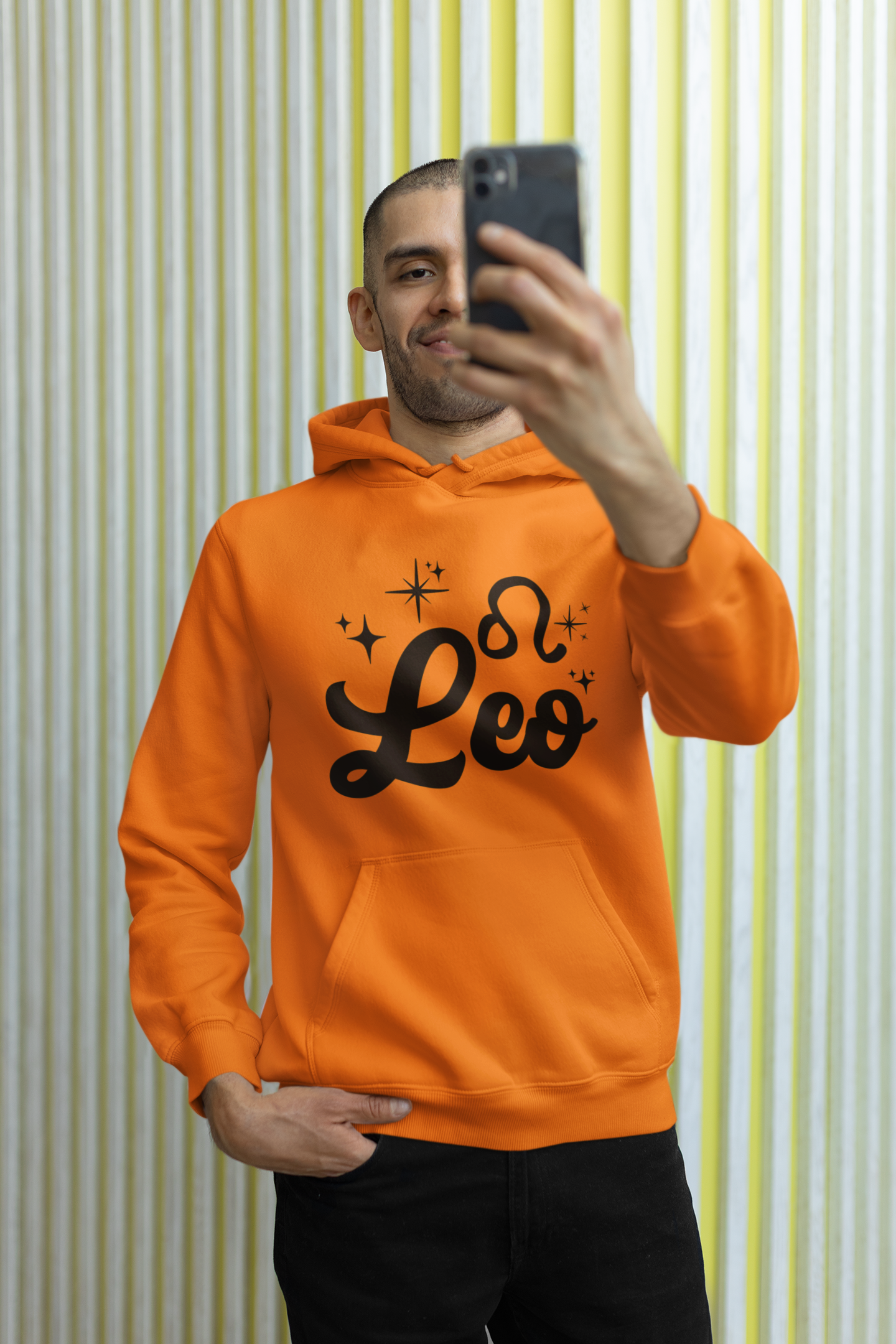 Person wearing a orange hoodie with 'Leo' black text, taking a selfie against a striped wall.