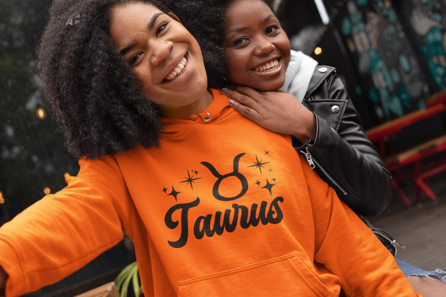 Two people posing together, one wearing a orange 'Taurus' hoodie, black text design 