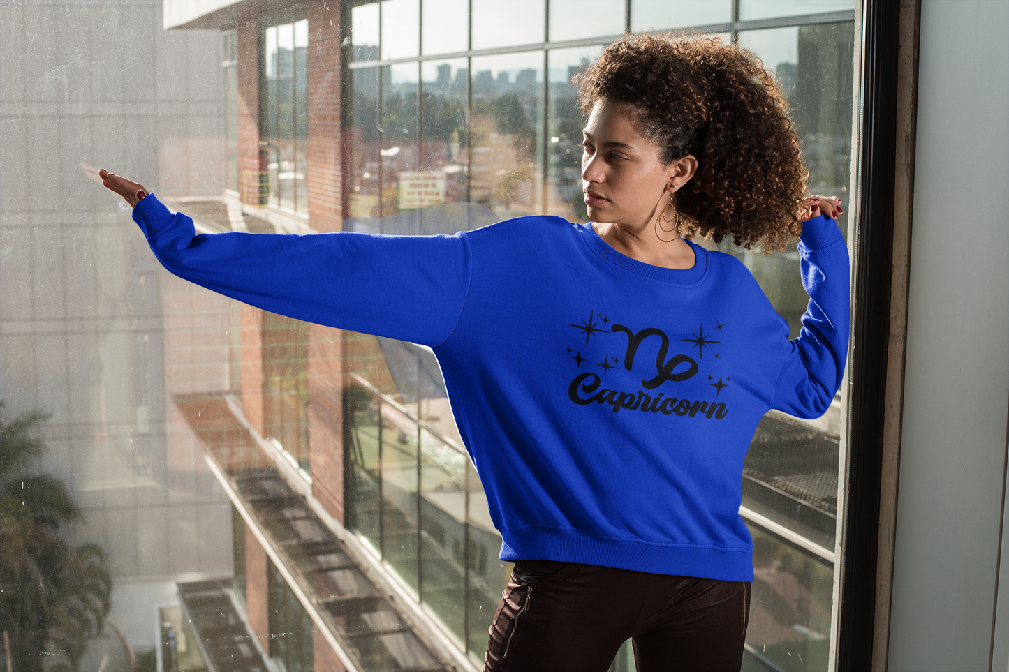Person wearing a blue 'Capricorn' black text design sweatshirt in front of a window with a cityscape view.