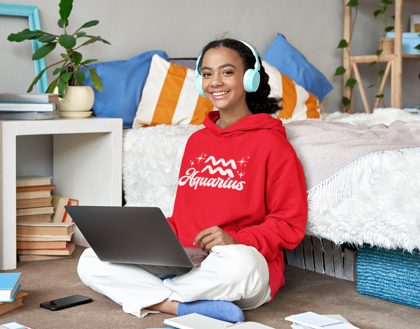 Person wearing a red hoodie 'Aquarius' white text with zodiac symbol, sitting on a bed with a laptop, surrounded by books and a plant.